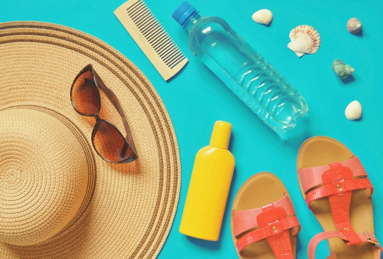 Sonnenhut, Sonnenbrille, Holzkamm, Mineralwasserflasche, Sonnencreme, Muschel und rote Sandalen.